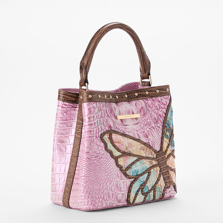 Brahmin pink satchel with textured embossed finish, brown handles and trim, large multicolored butterfly motif on the front, small gold-tone studs near top. Front view shows structured silhouette and top carry style.