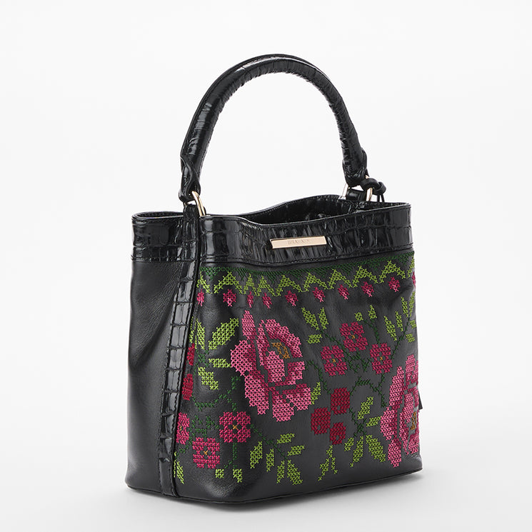Brahmin black satchel handbag with structured silhouette, textured handle and surface, gold-tone hardware, and vibrant pink and green floral cross-stitch pattern on the front; front exterior view shown.