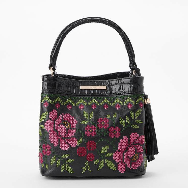 Brahmin black handbag with top handle, floral cross-stitch pattern in pink and green, black tassel accent on side, structured silhouette, and visible stitching. Front view showcasing bag’s texture and detailed design.