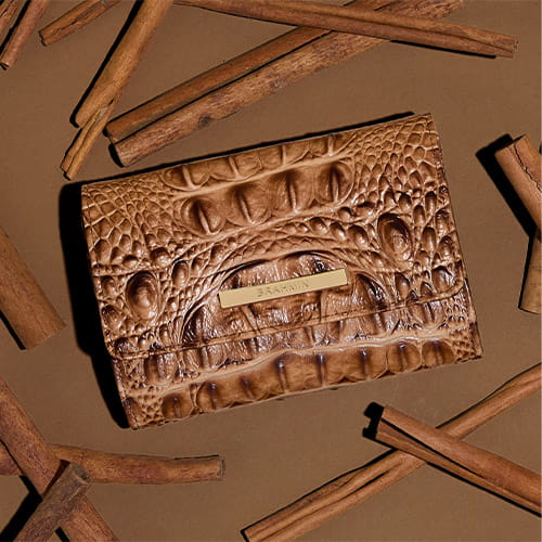 Brown crocodile-embossed leather wallet with gold logo bar, styled on a warm brown background with cinnamon sticks.