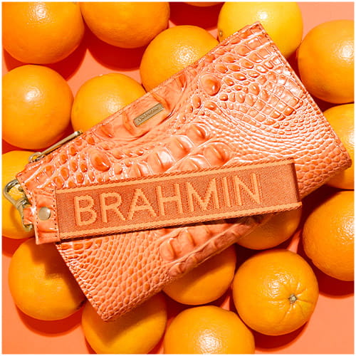 Orange crocodile-embossed leather wristlet with woven logo strap, styled on a background of fresh oranges.