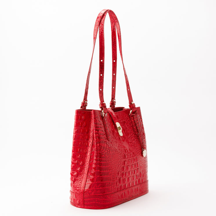 Side-profile view of a glossy, embossed red Fiora bucket bag by BRAHMIN, featuring two slender shoulder straps, gold-tone hardware, and a center snap closure.