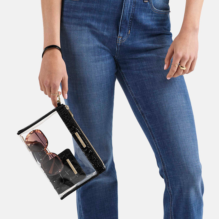 BRAHMIN Clarisse clear wristlet with black trim, showcased in hand. Transparent body reveals pink sunglasses, lipstick, and makeup brush inside. Black strap; silver hardware. Rings and a black bracelet are also visible.
