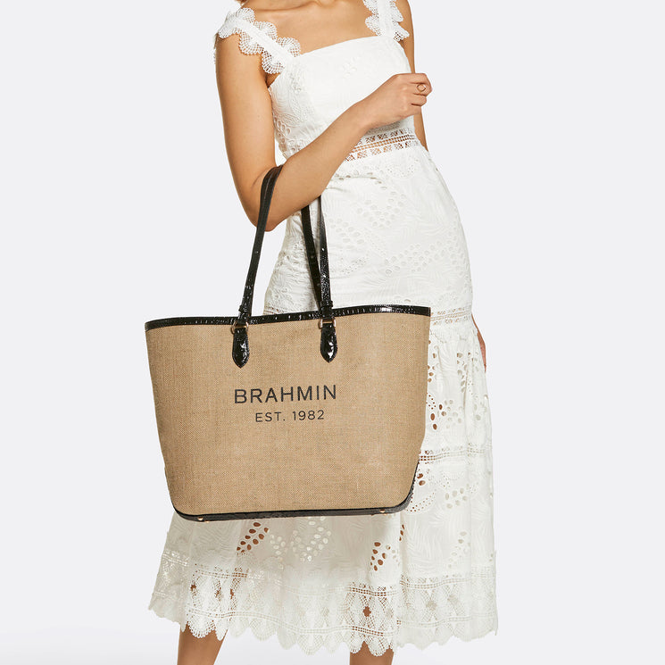 Brahmin beige tote bag with black straps, large silhouette, rectangular shape, and BRAHMIN EST. 1982 printed on the front. Carried on the arm by a woman in a white lace dress; face not visible.