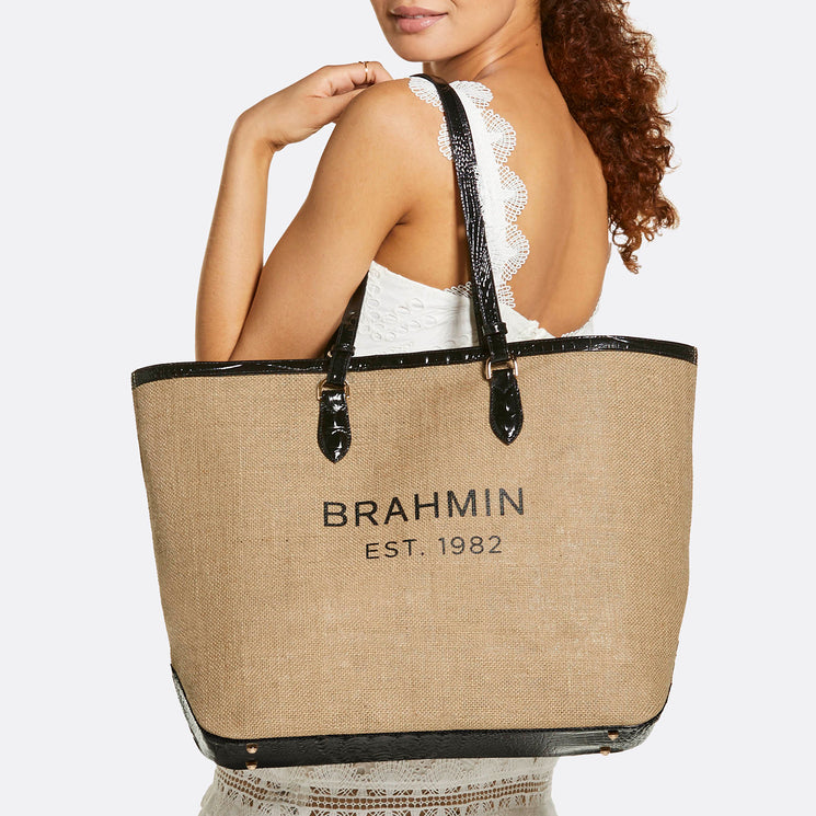 Brahmin large tan and black tote bag with BRAHMIN EST. 1982 printed on the front, slouchy silhouette, wide shoulder straps, carried over the shoulder by a woman in a white lace dress.