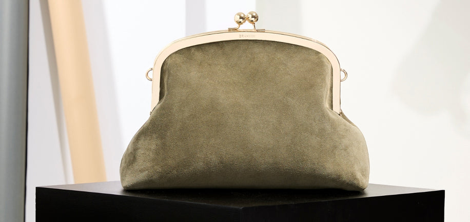 A muted olive green suede kiss-lock handbag with a gold-tone metal frame, displayed on a black pedestal against a clean, light background.