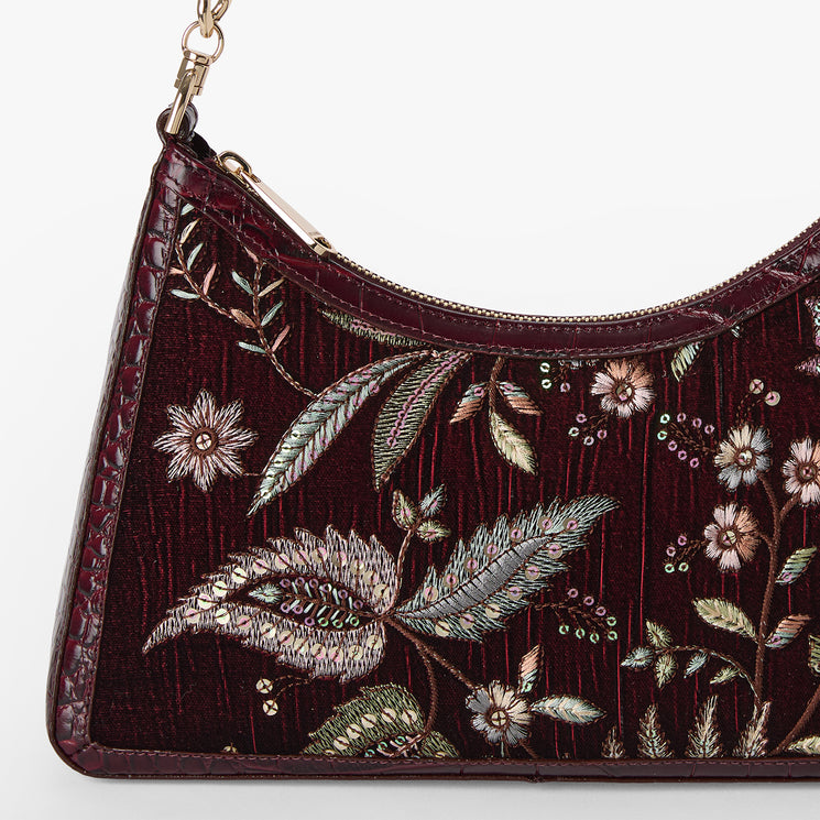 A close-up of a burgundy shoulder bag with intricate floral and leaf embroidery in metallic silver, gold, and green, featuring a textured crocodile-embossed side panel and a gold-tone chain strap.