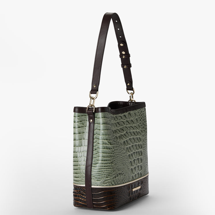 A structured shoulder bag with a sage green crocodile-embossed texture and dark brown leather trim, featuring a single adjustable shoulder strap with silver-tone studs, set against a plain light gray background.