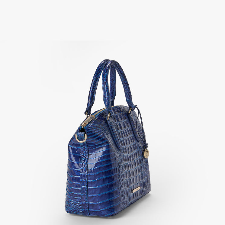 Brahmin blue embossed-texture leather satchel with short handles, gold-tone hardware, and a round gold charm, shown on a white background. The bag features a structured silhouette and elegant pattern detailing.
