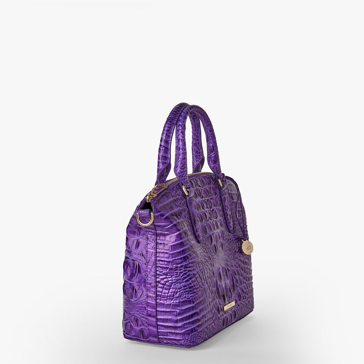 Brahmin purple embossed leather satchel with gold-tone hardware, dual rolled handles, structured silhouette, textured pattern, and displayed against a white background. No strap or interior is visible.