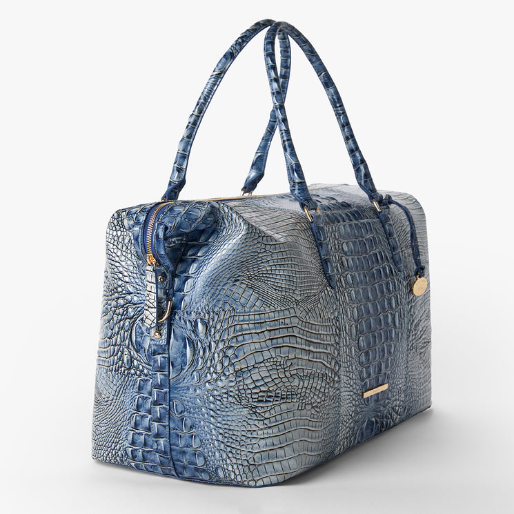 Brahmin large blue duffel bag with embossed textured pattern, two long handles, gold-tone hardware, zipper closure, and structured silhouette. Shown on a white background; interior not visible.