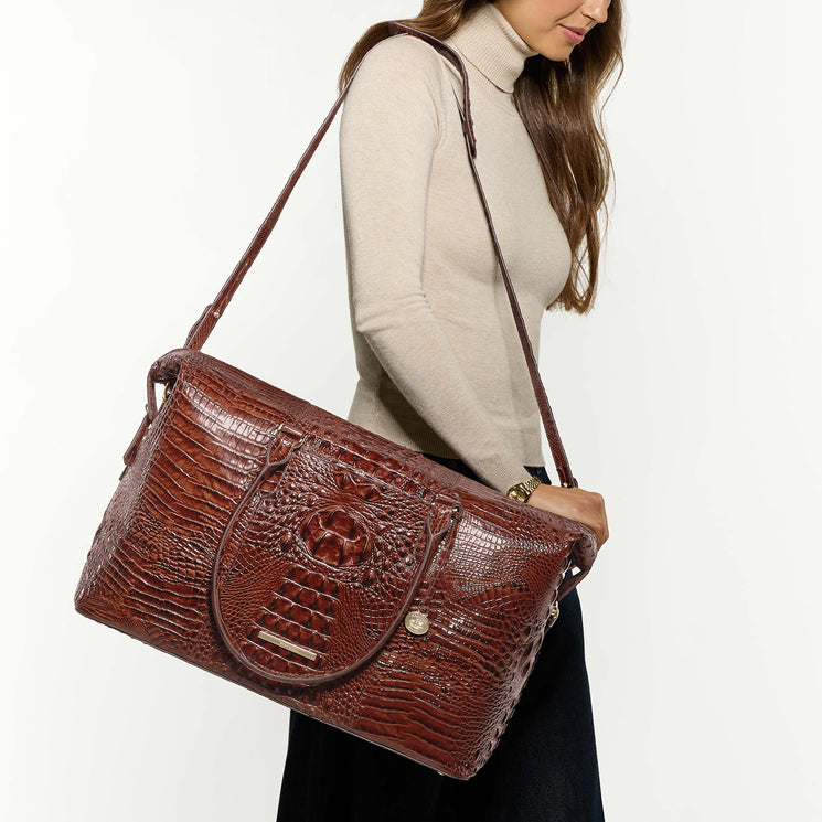 A woman wearing a beige turtleneck sweater and dark skirt carries a large brown crocodile-embossed shoulder bag against a plain white background.