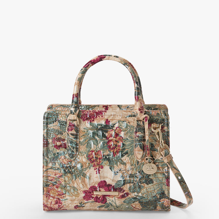 A structured handbag with a vintage floral tapestry pattern in muted red, green, and beige tones, featuring dual top handles, a detachable shoulder strap, and a hanging round metal charm.