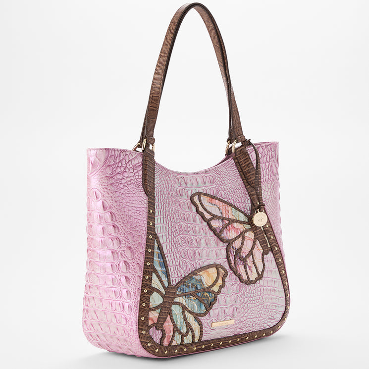 Brahmin pink satchel with embossed textured finish, brown rolled handles, gold-tone studs outlining the edges, and two colorful butterfly embellishments on the front; structured silhouette, front view.